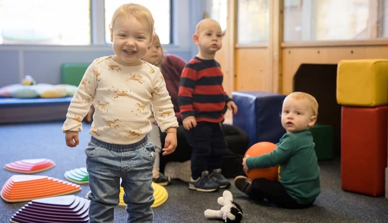 Children in the Older Infant Program playing with toys, one holding an orange ball in a brightly lit room.