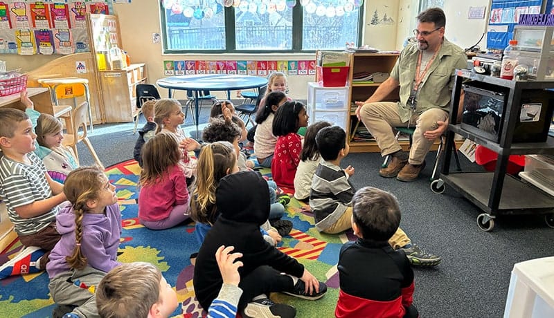 A classroom with young children sitting on a colorful rug, listening to a man speaking, next to a cart with a small animal enclosure.