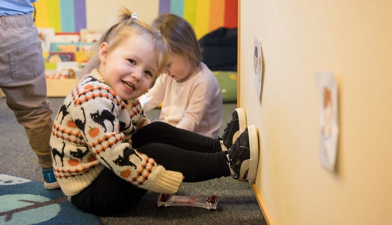 Young child in a colorful sweater smiles on the floor, with another child nearby and a rainbow wall hinting at early education.