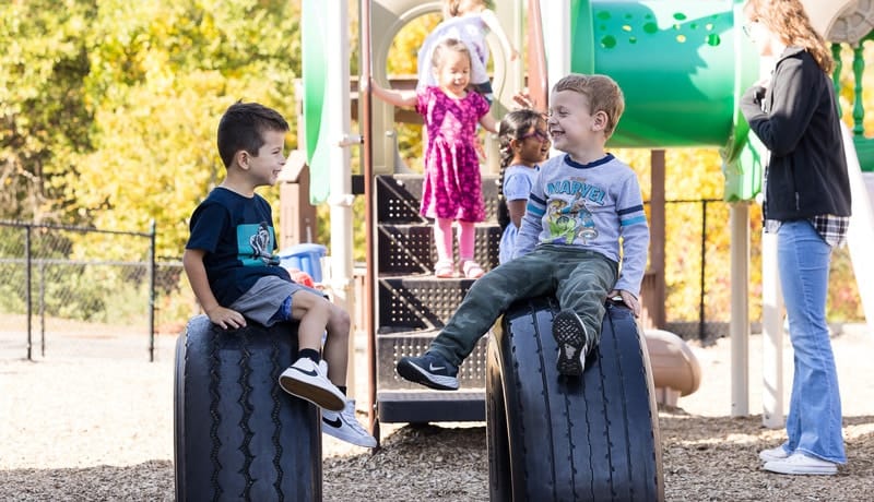 Two children sit on large tires in a playground, with other kids and an adult in the background near play equipment.
